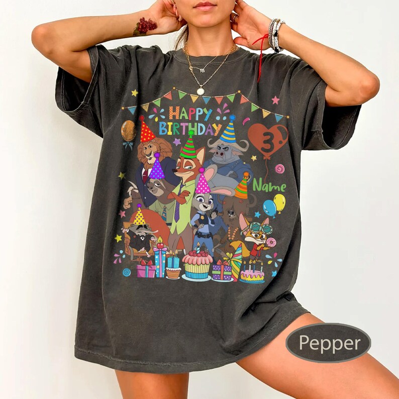 May include: A dark gray t-shirt featuring a colorful graphic of cartoon characters, party hats, and the text "Happy Birthday 3 Name". The design includes balloons, presents, and a cake. The shirt is a relaxed fit.