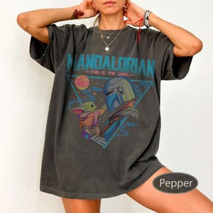 Pu&ograve; includere: Una t-shirt a maniche corte grigio antracite con una stampa grafica del Mandaloriano e Grogu. Il design include il testo "MANDALORIAN" e "THIS IS THE WAY" in verde acqua, con un pianeta rosa e una cornice triangolare blu.