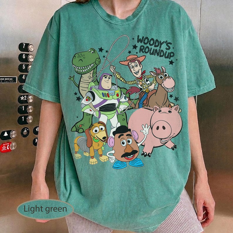 May include: Light green t-shirt featuring a graphic of characters from the movie Toy Story, including Woody, Buzz Lightyear, and Rex. The text "Woody's Roundup" is printed above the characters. The shirt has short sleeves and a relaxed fit.
