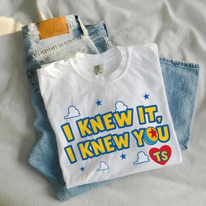 May include: A white short-sleeved t-shirt featuring the blue and yellow text I KNEW IT, I KNEW YOU with a red heart containing the letters TS. The design includes blue star and cloud outlines. The shirt is folded over a pair of light blue denim jeans.