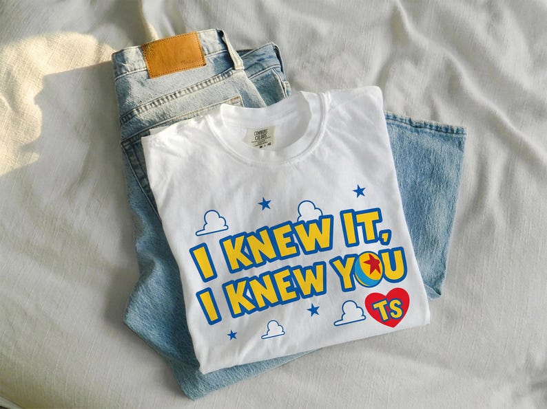 May include: A white short-sleeved t-shirt featuring the blue and yellow text I KNEW IT, I KNEW YOU with a red heart containing the letters TS. The design includes blue star and cloud outlines. The shirt is folded over a pair of light blue denim jeans.