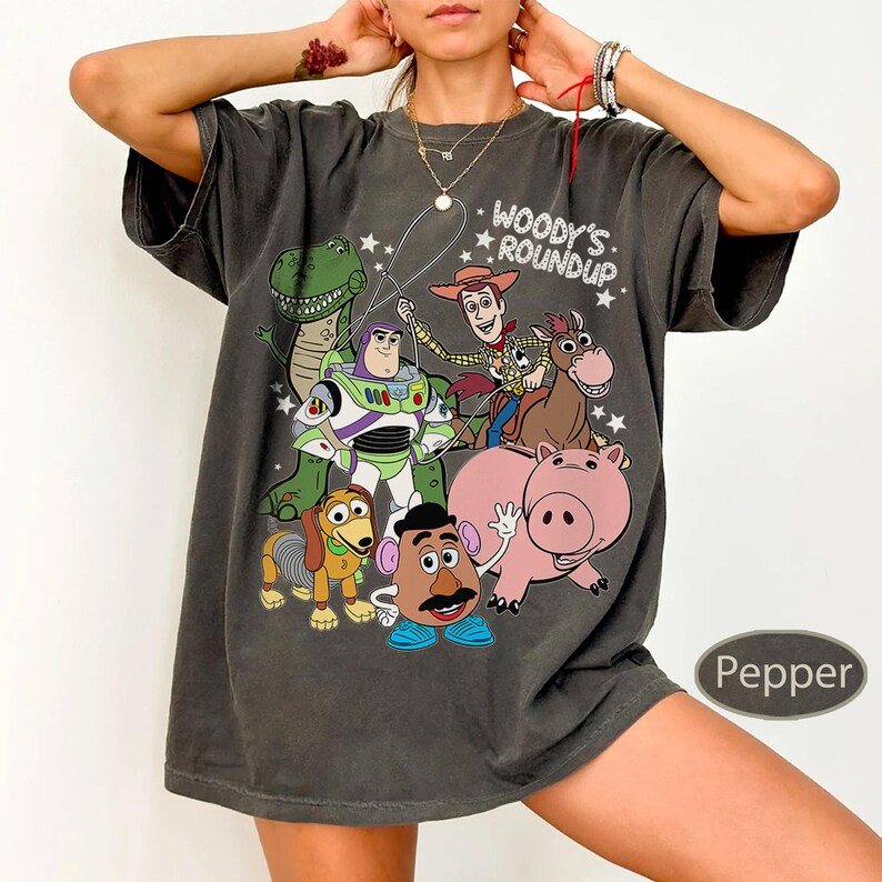 May include: Dark gray t-shirt featuring a colorful graphic of characters from the movie Toy Story, including Woody, Buzz Lightyear, and Rex. The text "Woody's Roundup" is above the characters. The shirt is a relaxed fit.