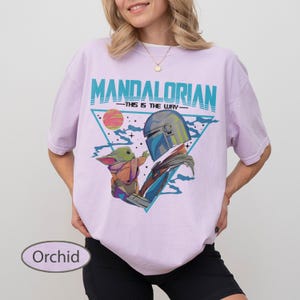 Pu&ograve; includere: Una t-shirt color orchidea con una stampa grafica del Mandaloriano e Grogu. Il design include il testo "MANDALORIAN" e "THIS IS THE WAY" in verde acqua, su uno sfondo triangolare con un pianeta e stelle in tonalit&agrave; di rosa e blu.
