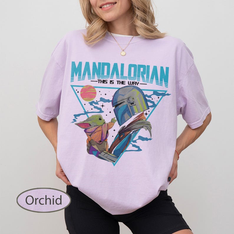 Pu&ograve; includere: Una t-shirt color orchidea con una stampa grafica del Mandaloriano e Grogu. Il design include il testo "MANDALORIAN" e "THIS IS THE WAY" in verde acqua, su uno sfondo triangolare con un pianeta e stelle in tonalit&agrave; di rosa e blu.