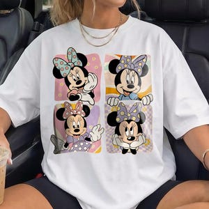 May include: White t-shirt featuring a colorful graphic of Minnie Mouse in four different poses. Each Minnie Mouse image is set against a unique background, including pink, orange, and purple hues. The shirt is a casual, oversized style.