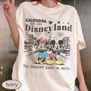 May include: Ivory-colored t-shirt featuring a graphic of Mickey and Minnie Mouse in front of Disneyland. The shirt includes the text "CALIFORNIA Disneyland EST. 1955" and "The happiest place on earth."