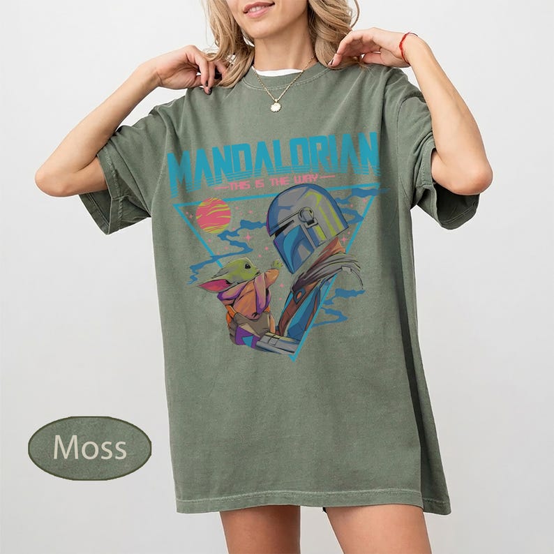 Pu&ograve; includere: Una t-shirt a maniche corte color verde muschio con una stampa grafica del Mandaloriano e Grogu. Il design include il testo "MANDALORIAN" e "THIS IS THE WAY" in blu, insieme a un pianeta stilizzato e accenti triangolari geometrici.