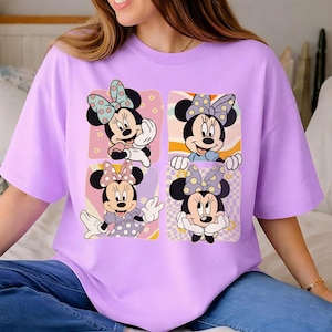 May include: A light purple t-shirt featuring a graphic of Minnie Mouse in four different poses. Each Minnie Mouse image has a polka dot bow and is set against a colorful background. The shirt is a casual, oversized style.