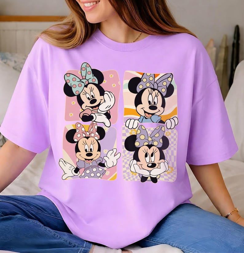 May include: A light purple t-shirt featuring a graphic of Minnie Mouse in four different poses. Each Minnie Mouse image has a polka dot bow and is set against a colorful background. The shirt is a casual, oversized style.