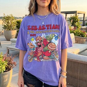 以下が含まれることがあります： 紫色の半袖Tシャツ。マラカスを演奏するカニのセバスチャンと他の海の生き物が描かれたグラフィックプリントが特徴です。テキストには「SEBASTIAN & HIS CRUSTACEAN BAND HOT」と書かれています。デザインにはサンゴ、泡、錨が含まれています。