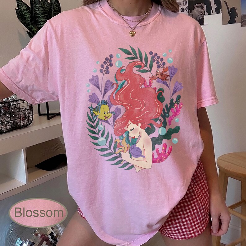 Peut inclure: T-shirt rose clair avec un motif color&eacute; d'une sir&egrave;ne aux cheveux roux, d'un poisson jaune et de plantes aquatiques. Le mot "Blossom" est imprim&eacute; en bas &agrave; gauche. Le t-shirt a des manches courtes et un col rond.