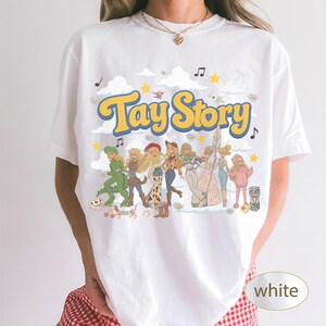 K&ouml;nnte beinhalten: Ein wei&szlig;es T-Shirt mit einem Grafikdruck, der den Text Tay Story in gelben und blauen Buchstaben zeigt. Das Design enth&auml;lt mehrere illustrierte Figuren in verschiedenen Outfits, umgeben von Wolken, Sternen und Musiknoten auf wei&szlig;em Hintergrund.