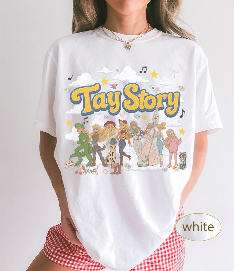 K&ouml;nnte beinhalten: Ein wei&szlig;es T-Shirt mit einem Grafikdruck, der den Text Tay Story in gelben und blauen Buchstaben zeigt. Das Design enth&auml;lt mehrere illustrierte Figuren in verschiedenen Outfits, umgeben von Wolken, Sternen und Musiknoten auf wei&szlig;em Hintergrund.