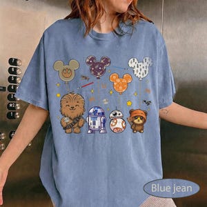 May include: Blue jean colored t-shirt featuring cartoon characters and Mickey Mouse-shaped balloons. The design includes Chewbacca, R2-D2, BB-8, and an Ewok. The balloons are in various colors and patterns, with stars and lightsabers.