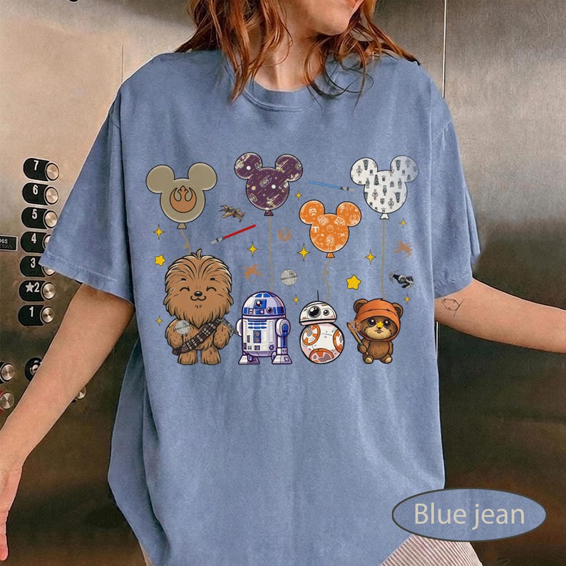 May include: Blue jean colored t-shirt featuring cartoon characters and Mickey Mouse-shaped balloons. The design includes Chewbacca, R2-D2, BB-8, and an Ewok. The balloons are in various colors and patterns, with stars and lightsabers.