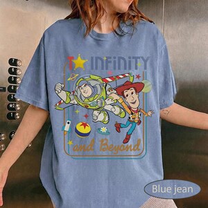 May include: Blue jean short-sleeve t-shirt featuring Buzz Lightyear and Woody from Toy Story. The shirt has the text "Infinity and Beyond" with a star and rocket ship graphic. The shirt is a light blue color.