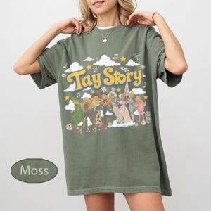 K&ouml;nnte beinhalten: Ein moosgr&uuml;nes Kurzarm-T-Shirt mit einem Grafikdruck, der den Text Tay Story in gelben Buchstaben zeigt. Das Design enth&auml;lt verschiedene illustrierte Figuren, wei&szlig;e Wolken, gelbe Sterne und Noten, die auf der Vorderseite verteilt sind.