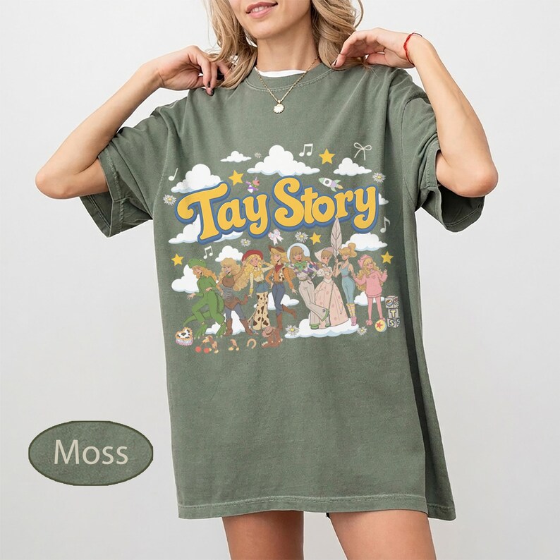 K&ouml;nnte beinhalten: Ein moosgr&uuml;nes Kurzarm-T-Shirt mit einem Grafikdruck, der den Text Tay Story in gelben Buchstaben zeigt. Das Design enth&auml;lt verschiedene illustrierte Figuren, wei&szlig;e Wolken, gelbe Sterne und Noten, die auf der Vorderseite verteilt sind.