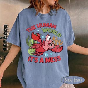 May include: Blue jean colored t-shirt featuring a cartoon crab with the text "THE HUMAN WORLD IT'S A MESS". The crab is red and pink, with green seaweed and coral accents. The shirt is a casual, short-sleeved style.