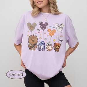 May include: Orchid-colored t-shirt featuring a graphic of Star Wars characters and Mickey Mouse-shaped balloons. The design includes Chewbacca, R2-D2, BB-8, and an Ewok. The shirt has a relaxed fit.
