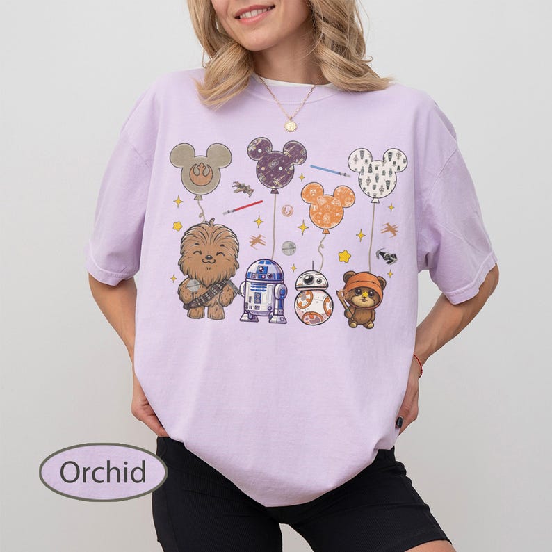 May include: Orchid-colored t-shirt featuring a graphic of Star Wars characters and Mickey Mouse-shaped balloons. The design includes Chewbacca, R2-D2, BB-8, and an Ewok. The shirt has a relaxed fit.