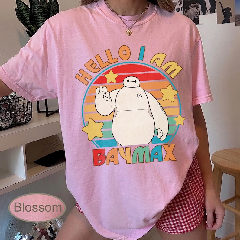 May include: Pink t-shirt featuring a graphic of Baymax from Big Hero 6. The design includes the text "HELLO I AM BAYMAX" in yellow and red, with a circular background of orange, blue, and yellow stripes and stars.