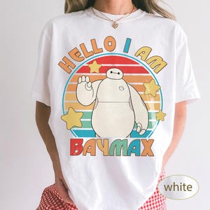 May include: White t-shirt featuring a cartoon image of Baymax with the text "HELLO I AM BAYMAX" in orange, red, and blue. The design includes a circular background with yellow stars and a striped sunset design.