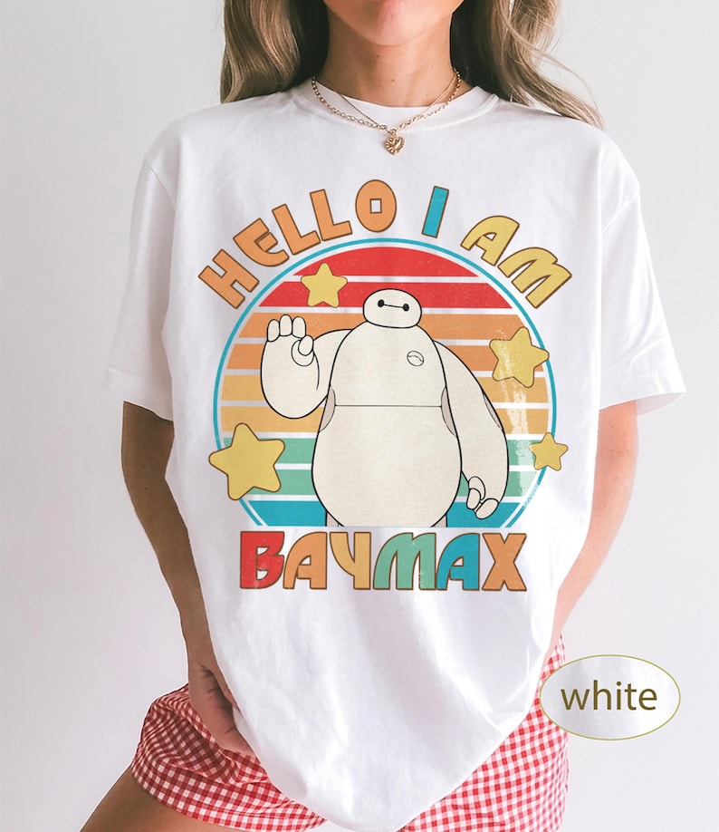 May include: White t-shirt featuring a cartoon image of Baymax with the text "HELLO I AM BAYMAX" in orange, red, and blue. The design includes a circular background with yellow stars and a striped sunset design.