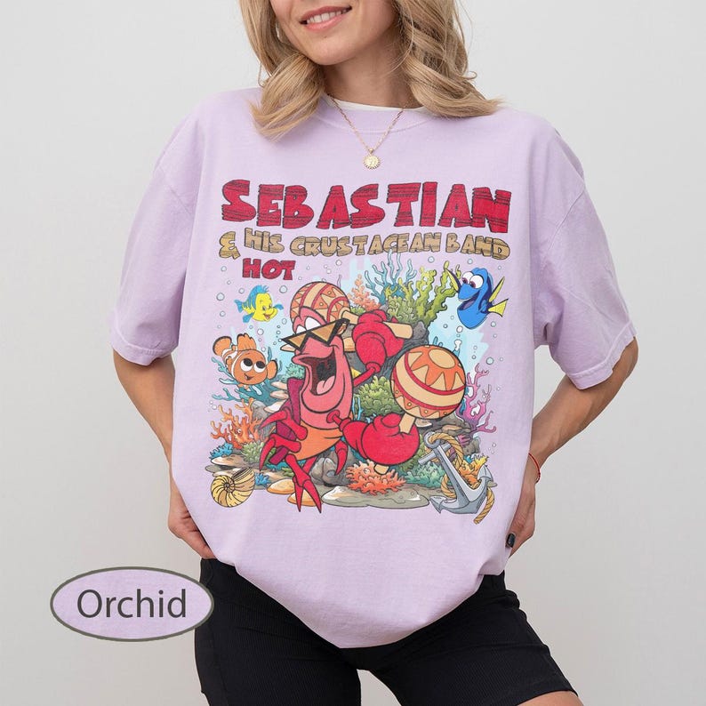 以下が含まれることがあります： オーキッドカラーのTシャツで、マラカスを演奏するカニのセバスチャンと他の海の生き物が描かれたグラフィックプリントが特徴です。テキストには「SEBASTIAN & HIS CRUSTACEAN BAND HOT」と書かれています。デザインにはサンゴ、錨、カラフルな魚が含まれています。