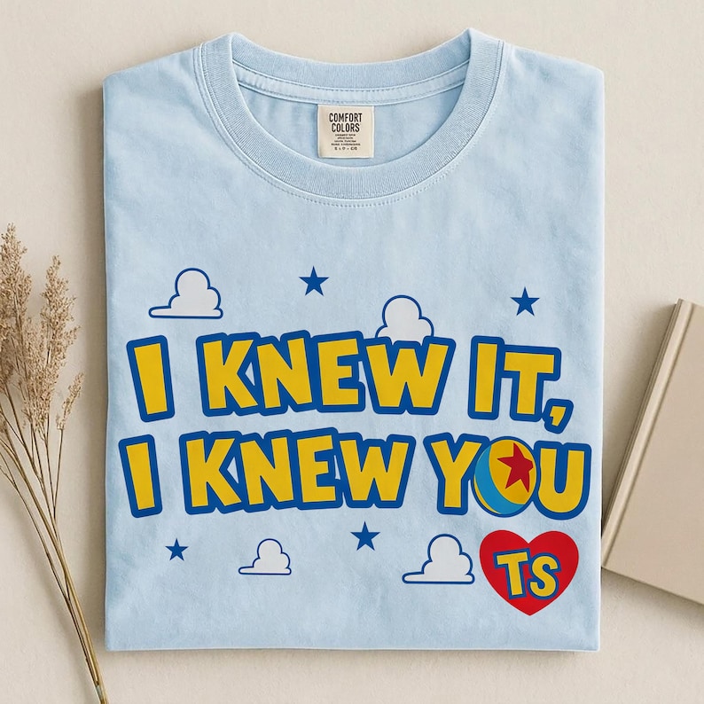 May include: A light blue crew neck t-shirt featuring the yellow text I KNEW IT, I KNEW YOU with a red heart containing the letters TS and a small ball graphic. The design includes white cloud outlines and dark blue stars printed on the front.
