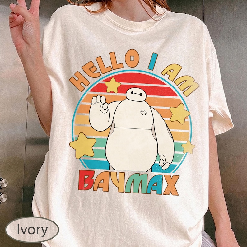 May include: Ivory-colored t-shirt featuring a graphic of Baymax from Big Hero 6. The design includes the text "HELLO I AM BAYMAX" in colorful lettering, with a circular background and star accents. The shirt has short sleeves.