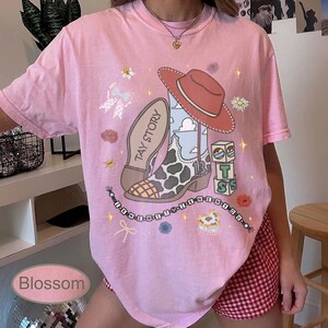 May include: A light pink short-sleeve t-shirt featuring a graphic print of a cowboy boot with a cow print pattern, a red cowboy hat, a stack of blocks, and a beaded necklace design with the text I KNEW IT I KNEW YOU and TAY STORY.