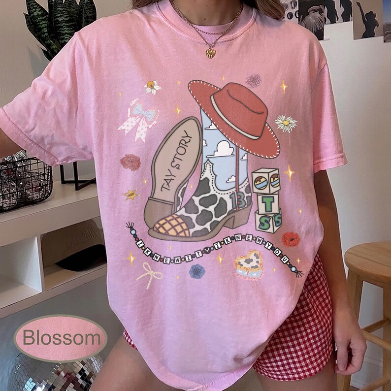 May include: A light pink short-sleeve t-shirt featuring a graphic print of a cowboy boot with a cow print pattern, a red cowboy hat, a stack of blocks, and a beaded necklace design with the text I KNEW IT I KNEW YOU and TAY STORY.