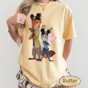 Pu&ograve; includere: T-shirt color burro con un'illustrazione di Nick Wilde e Judy Hopps di Zootropolis. Nick, una volpe, indossa un cappello e Judy, un coniglio, tiene lo zucchero filato. La parola "Butter" &egrave; stampata in basso a destra.