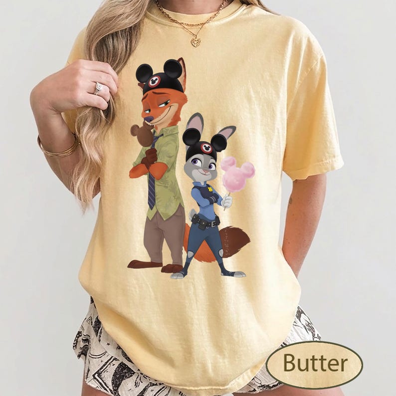 Pu&ograve; includere: T-shirt color burro con un'illustrazione di Nick Wilde e Judy Hopps di Zootropolis. Nick, una volpe, indossa un cappello e Judy, un coniglio, tiene lo zucchero filato. La parola "Butter" &egrave; stampata in basso a destra.