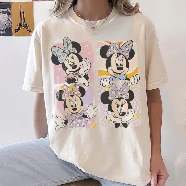 May include: Cream-colored t-shirt featuring a Minnie Mouse graphic. The graphic showcases four different Minnie Mouse poses, each within a pastel-colored square. Minnie is wearing polka-dot bows and dresses. The shirt has short sleeves and a crew neck.