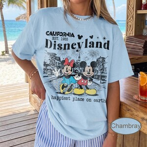 May include: Light blue t-shirt with a graphic of Mickey and Minnie Mouse in front of Disneyland. The shirt has the text "CALIFORNIA EST. 1955 Disneyland happiest place on earth" in black lettering. The shirt is a casual style.