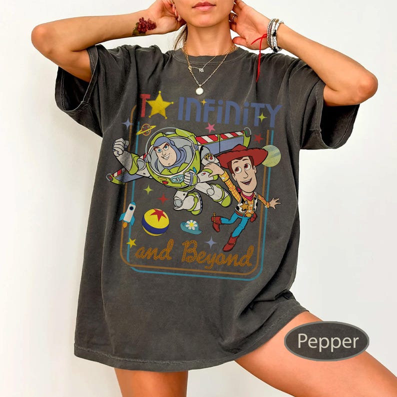 May include: Dark gray t-shirt featuring Buzz Lightyear and Woody from Toy Story, with the text "To Infinity and Beyond." The design includes a star, rocket, and planet details. The shirt is a relaxed fit.