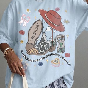 May include: A light blue t-shirt featuring a graphic print of a red cowboy hat, a cow-print boot with the text TAY STORY, a stack of blocks with the letters T, S, and the number 13, and a beaded string with the text I KNEW IT WAS YOU.