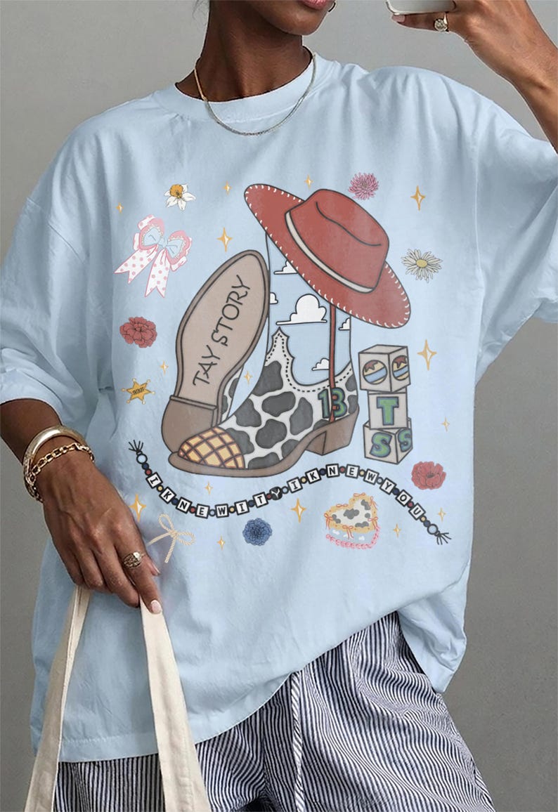 May include: A light blue t-shirt featuring a graphic print of a red cowboy hat, a cow-print boot with the text TAY STORY, a stack of blocks with the letters T, S, and the number 13, and a beaded string with the text I KNEW IT WAS YOU.