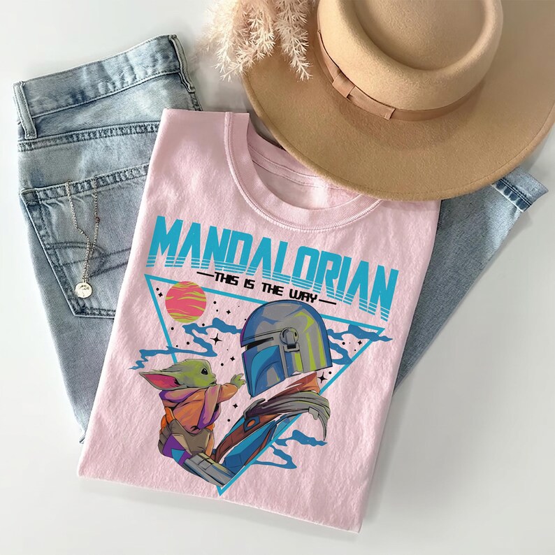 Pu&ograve; includere: Una t-shirt rosa chiaro con una stampa grafica di The Mandalorian e Grogu. Il testo recita MANDALORIAN THIS IS THE WAY. Il design include un triangolo blu, un pianeta e delle stelle. La maglietta &egrave; abbinata a jeans azzurri e un cappello beige.
