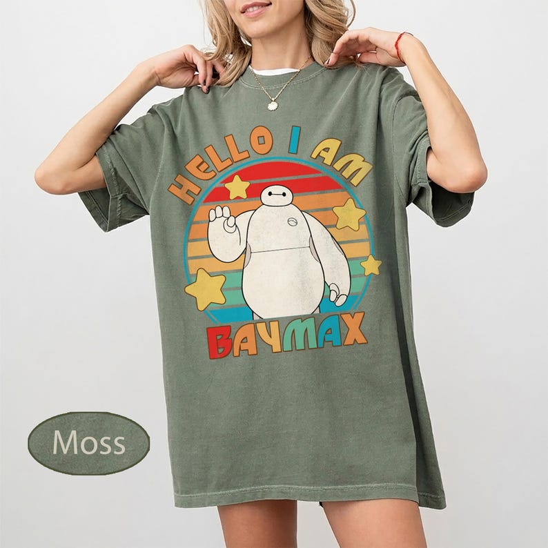 May include: Moss green t-shirt featuring a graphic of Baymax from Big Hero 6. The design includes the text "HELLO I AM BAYMAX" in a retro style with a circular background and stars.