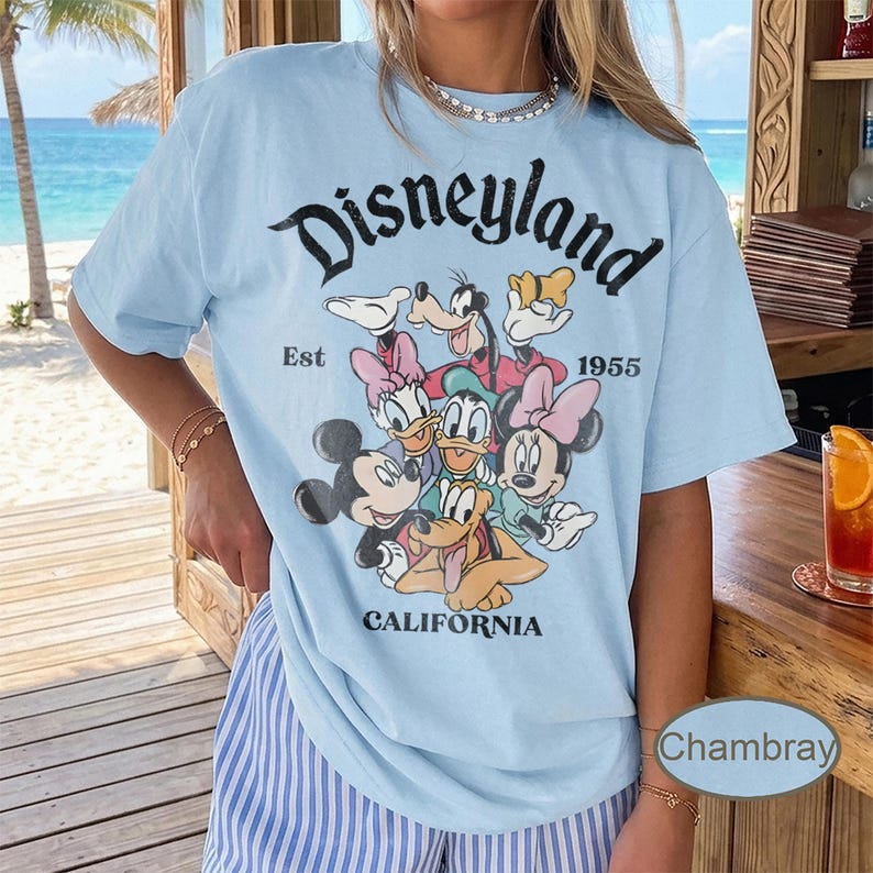 May include: Light blue t-shirt featuring a graphic of Disney characters, including Mickey and Minnie Mouse, Donald and Daisy Duck, and Goofy. The word "Disneyland" is arched across the top, with "Est 1955" and "California" below.