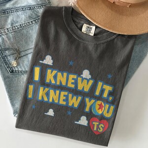 May include: A charcoal gray short-sleeve t-shirt featuring the yellow text "I KNEW IT, I KNEW YOU" with blue outlines. The design includes white cloud shapes, blue stars, a colorful ball with a red star, and a red heart containing the yellow letters "TS".