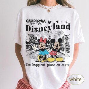 May include: White t-shirt featuring a graphic of Mickey and Minnie Mouse in front of a Disneyland scene. The shirt has the text "CALIFORNIA EST. 1955 Disneyland" and "The happiest place on earth".