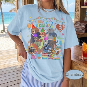 May include: Light blue t-shirt featuring a colorful graphic of cartoon animals celebrating a birthday. The design includes the text "Happy Birthday" and a number 3 balloon. The shirt is decorated with balloons, presents, and a cupcake.
