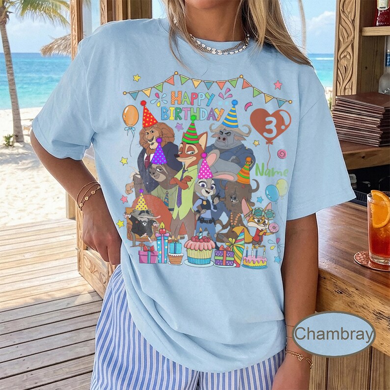 May include: Light blue t-shirt featuring a colorful graphic of cartoon animals celebrating a birthday. The design includes the text "Happy Birthday" and a number 3 balloon. The shirt is decorated with balloons, presents, and a cupcake.