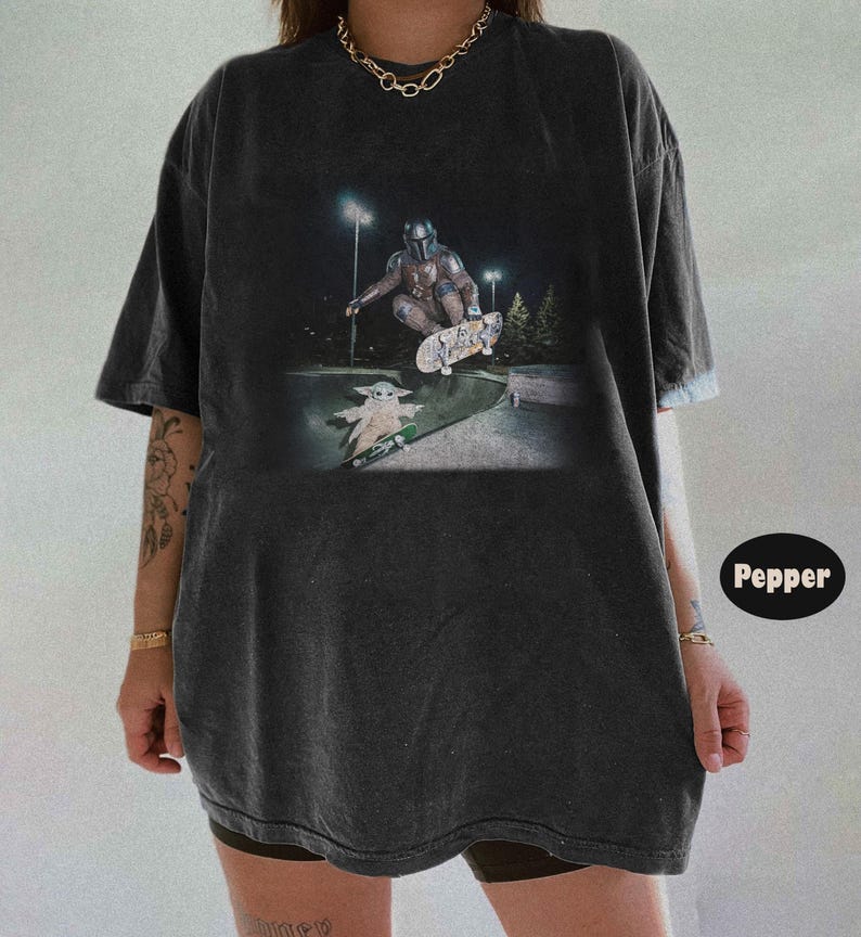 May include: A charcoal gray oversized t-shirt featuring a graphic print of a character in armor skateboarding in the air above a small creature on a skateboard. The shirt has short sleeves, a crew neckline, and a faded, washed-out fabric texture.