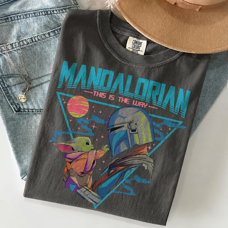 Pu&ograve; includere: Una t-shirt grigio antracite con una stampa grafica di The Mandalorian e Grogu. Il design include il testo "MANDALORIAN" e "THIS IS THE WAY" in blu brillante, un pianeta giallo e un triangolo blu. La maglietta ha un'etichetta Comfort Colors.