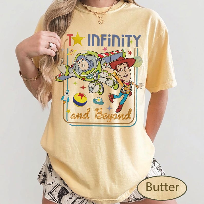 May include: Butter-colored t-shirt featuring a graphic of Buzz Lightyear and Woody from Toy Story, with the text "To Infinity and Beyond." The design includes a star, rocket, and other playful elements. The shirt is a relaxed fit.